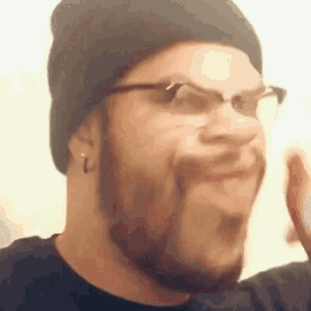 a man with a beard and glasses is wearing a black hat and making a funny face .