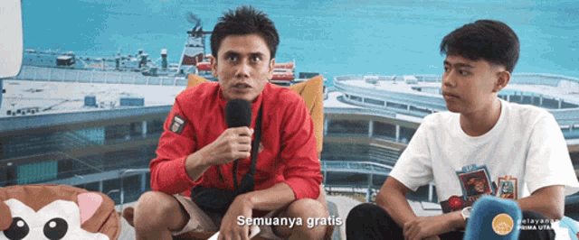 a man in a red jacket is talking into a microphone while another man sits next to him and says " semuanya gratis "