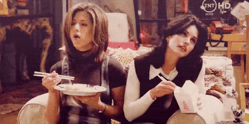 two women are sitting next to each other on a couch eating food with chopsticks .