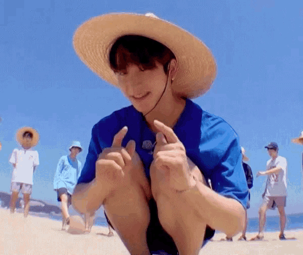 a young man wearing a blue shirt and a straw hat is squatting on the beach