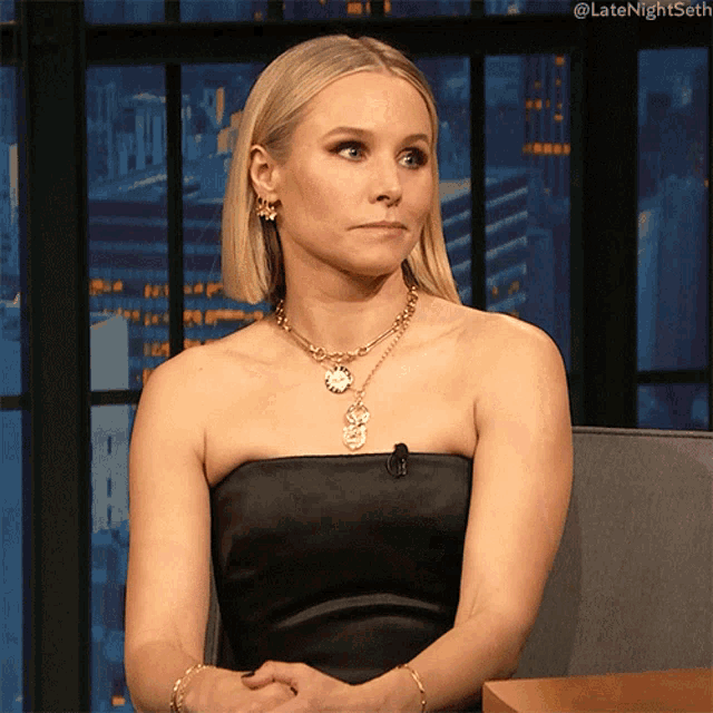 a woman wearing a black strapless dress and a gold necklace is sitting in a chair