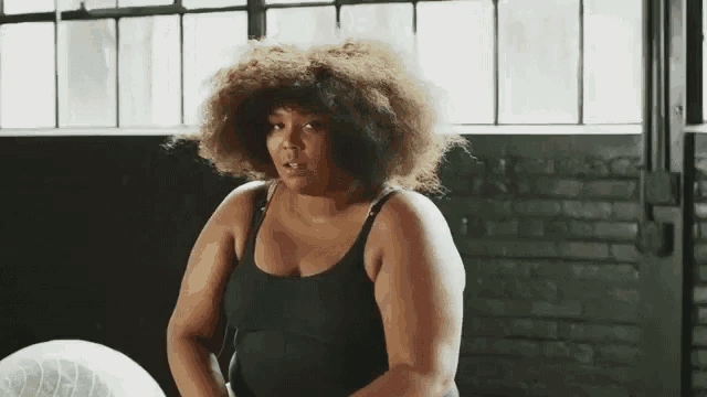 a woman with curly hair and a black tank top is sitting in front of a fan .
