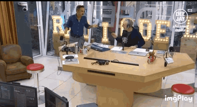 two men are sitting at a table in front of a sign that says deejay chiama italia