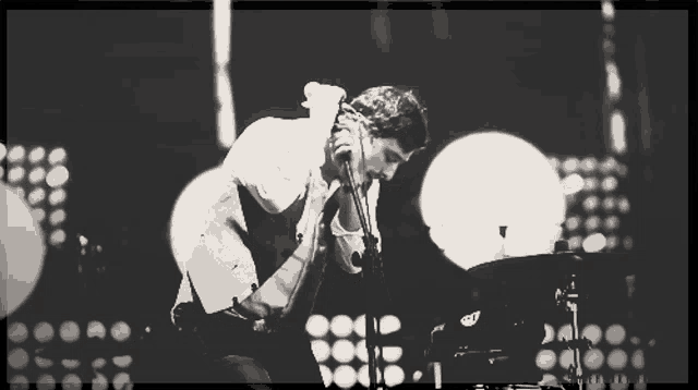 a black and white photo of a man singing into a microphone with a drum set behind him