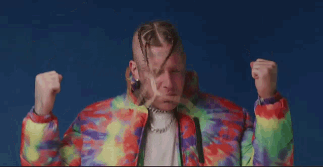 a man wearing a tie dye jacket holds his hands up in the air