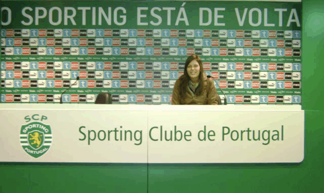 a woman sits at a desk in front of a wall that says o sporting esta de volta