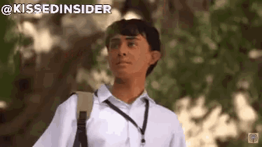 a boy in a white shirt with a black lanyard around his neck stands in front of trees with the words " kissedinsider " above him