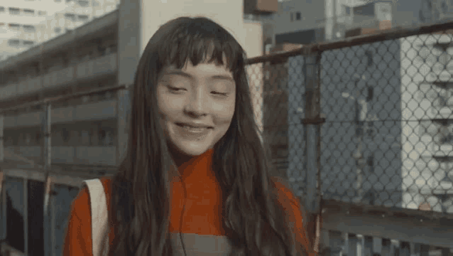 a woman with long hair and bangs is smiling with her eyes closed in front of a chain link fence .