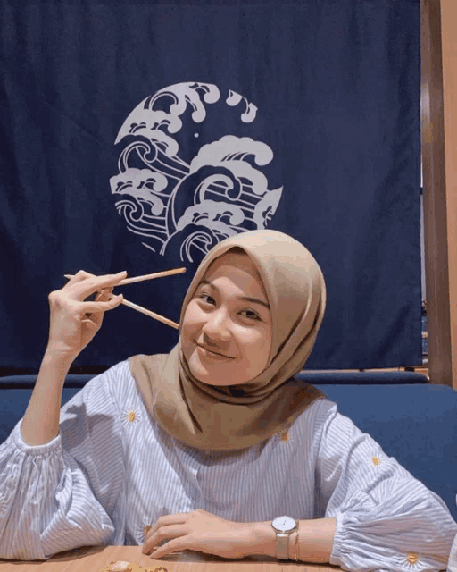 a woman in a hijab is eating with chopsticks in front of a blue curtain with waves on it