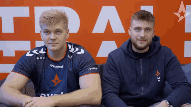 a man wearing a hummel shirt sits next to another man wearing a hoodie