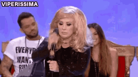 a woman in a wig is talking into a microphone while a man and a woman watch .