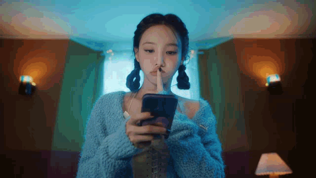 a woman in a blue sweater is holding her finger to her lips while looking at her cell phone .