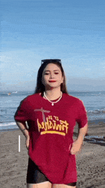 a woman is standing on a beach wearing a red t-shirt with a cross on it .