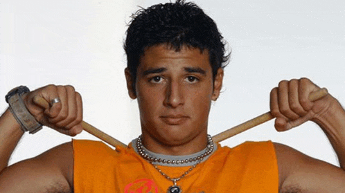 a man wearing an orange tank top and a necklace is holding two sticks over his shoulder