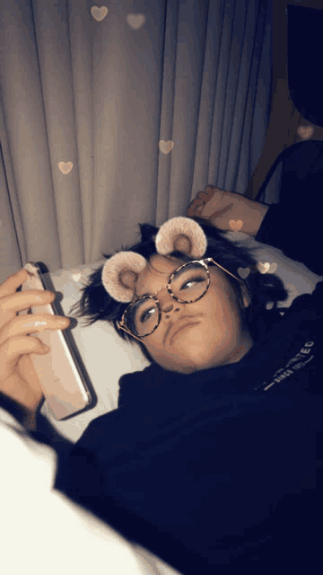 a person wearing glasses and bear ears laying on a bed looking at their phone