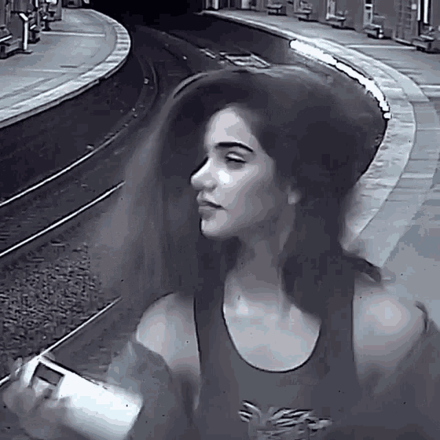 a woman wearing a black tank top with a bird on it is standing next to train tracks