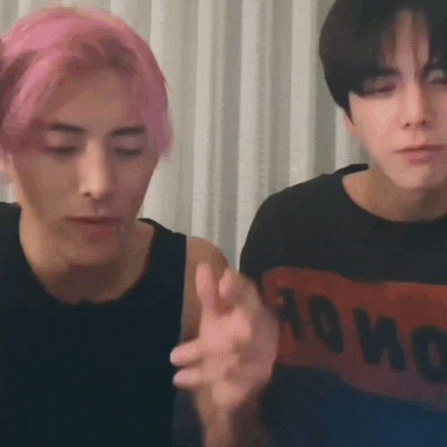 two young men are sitting next to each other in a room . one of the men has pink hair .