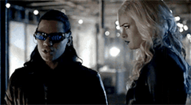 a man and a woman are standing next to each other in a dark room . the woman is wearing sunglasses .