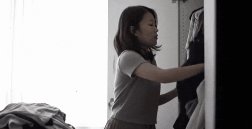a woman in a grey shirt is standing in a closet looking at clothes