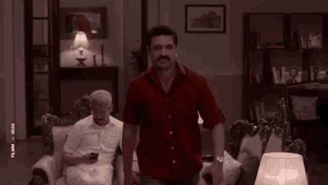 a man in a red shirt is standing in a living room next to a man in a white shirt sitting in a chair .