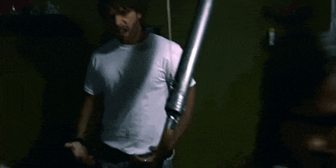 a man in a white t-shirt is standing in a dark room next to a metal pipe .