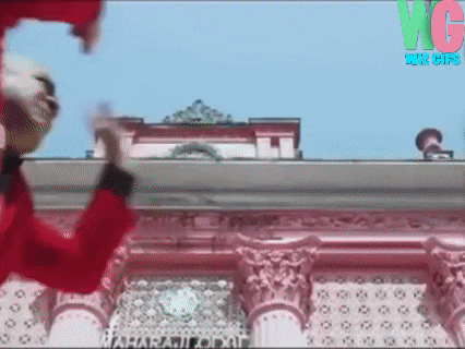 a man in a red jacket is jumping in the air in front of a building with the letters vg on it .
