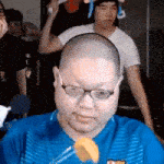 a bald man wearing glasses and a blue shirt is sitting in front of a crowd .