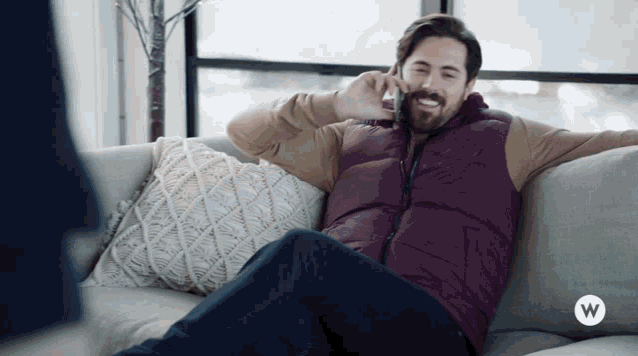 a man sits on a couch talking on a cell phone with a white w on the bottom right