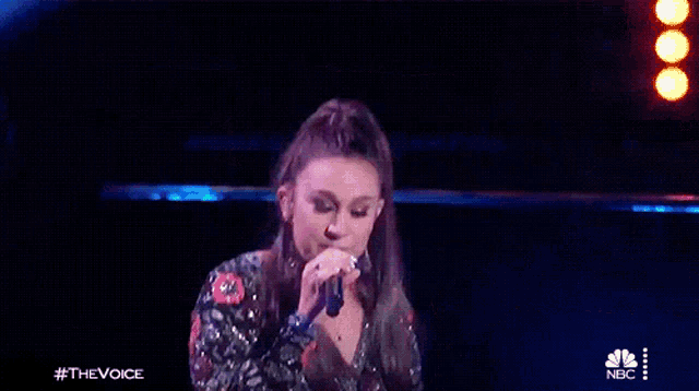a woman is singing into a microphone on a stage with a nbc logo in the background