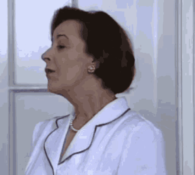 a woman in a white coat and pearls is standing next to a door .