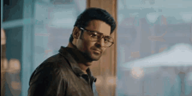 a man wearing glasses and a leather jacket is standing in front of a window looking at the camera .