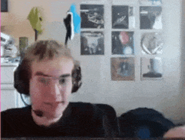 a man wearing headphones and glasses looks at the camera in front of a wall with pictures on it