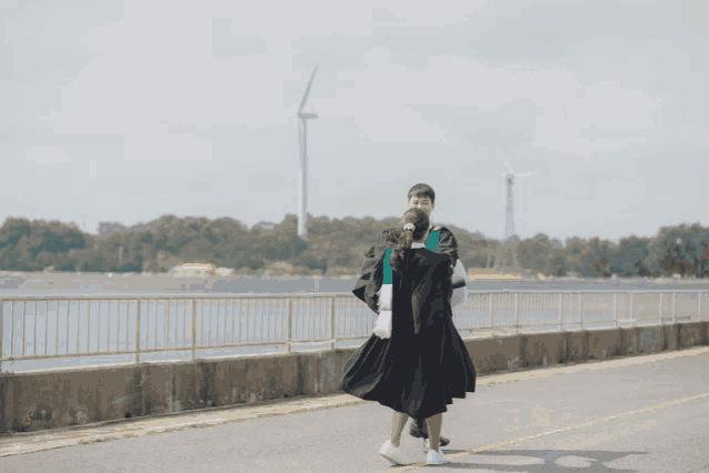 a man and a woman in graduation gowns are walking by a body of water with a windmill in the background