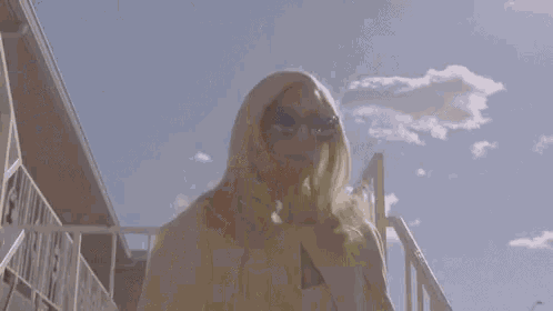 a blonde woman wearing sunglasses and a yellow cape is standing on a balcony .