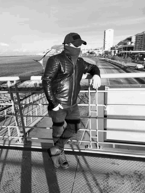a man wearing a leather jacket and a black hat stands on a bridge overlooking the ocean