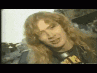 a close up of a man with long blonde hair wearing a black shirt with the letter m on it .