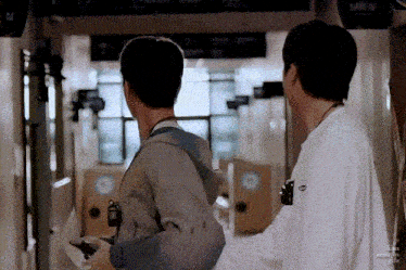 two men are standing next to each other in a hallway . one of the men is holding a camera .