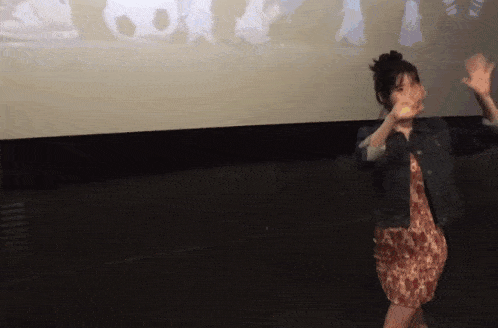 a woman in a dress and denim jacket is dancing in front of a screen