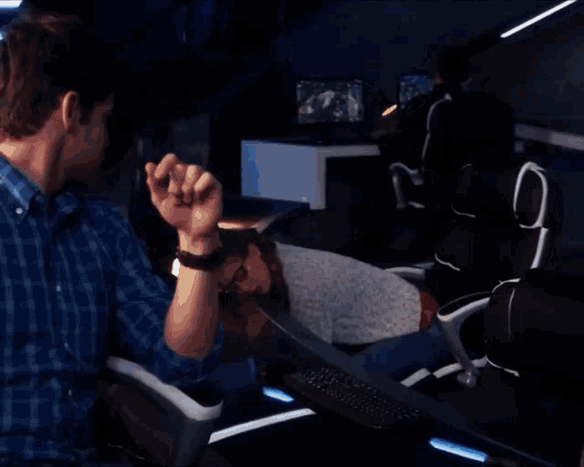 a man in a blue plaid shirt is playing a video game with a woman laying on the floor behind him