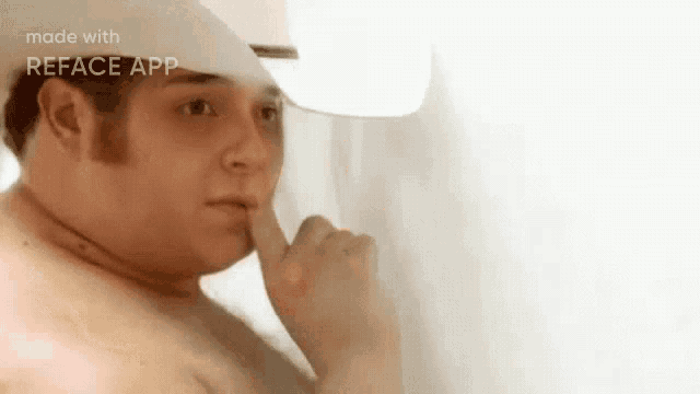 a man without a shirt is looking out a window with his finger to his mouth .