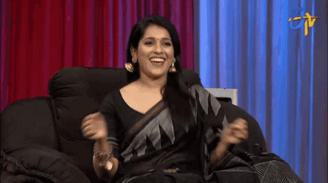 a woman in a black and grey saree is sitting on a couch and smiling .