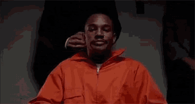a man in an orange prison uniform is sitting in a chair with a man holding his ear .