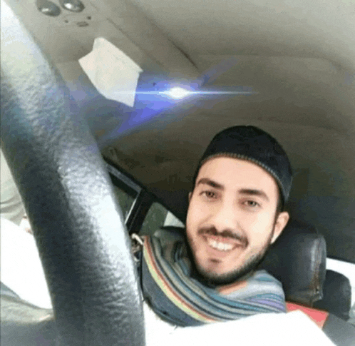 a man wearing a hat and scarf smiles while sitting in the driver 's seat of a car