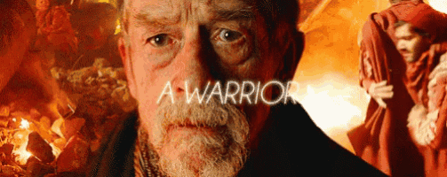 a man with a beard is standing in front of a fire with the words a warrior on his face .