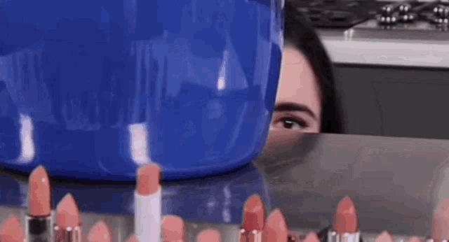 a woman is peeking out from behind a table full of lipsticks and a blue bucket .
