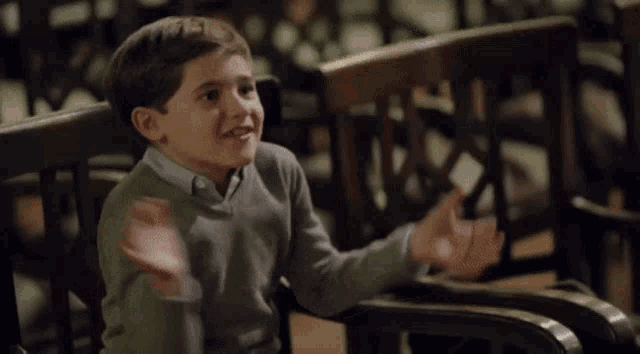 a young boy in a grey sweater is sitting on a bench with his hands outstretched