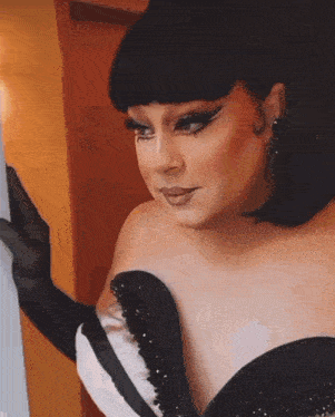 a drag queen in a black and white dress is looking at herself in a mirror