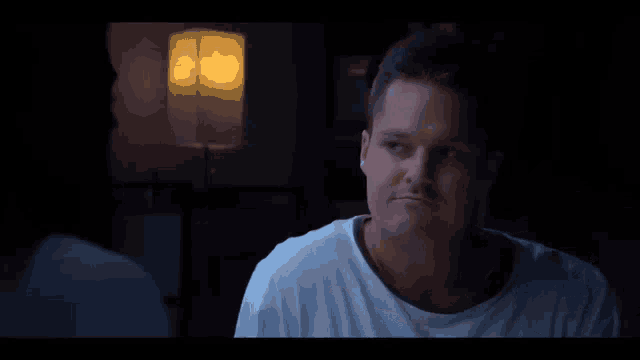 a man in a white shirt is sitting in front of a lamp in a dark room .