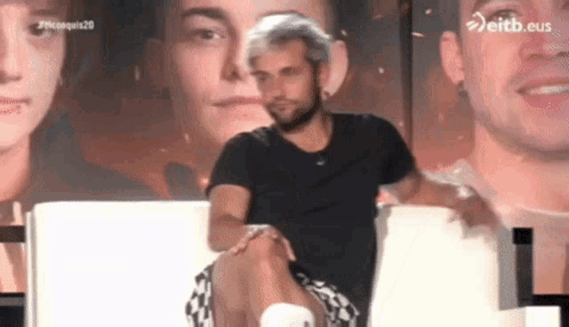 a man in a black shirt is sitting on a white chair with his legs crossed in front of a picture of a woman .