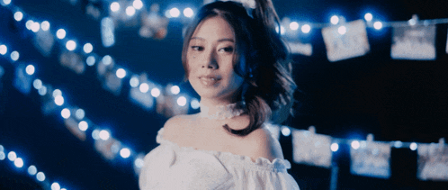 a woman wearing a white off the shoulder top stands in front of a string of lights